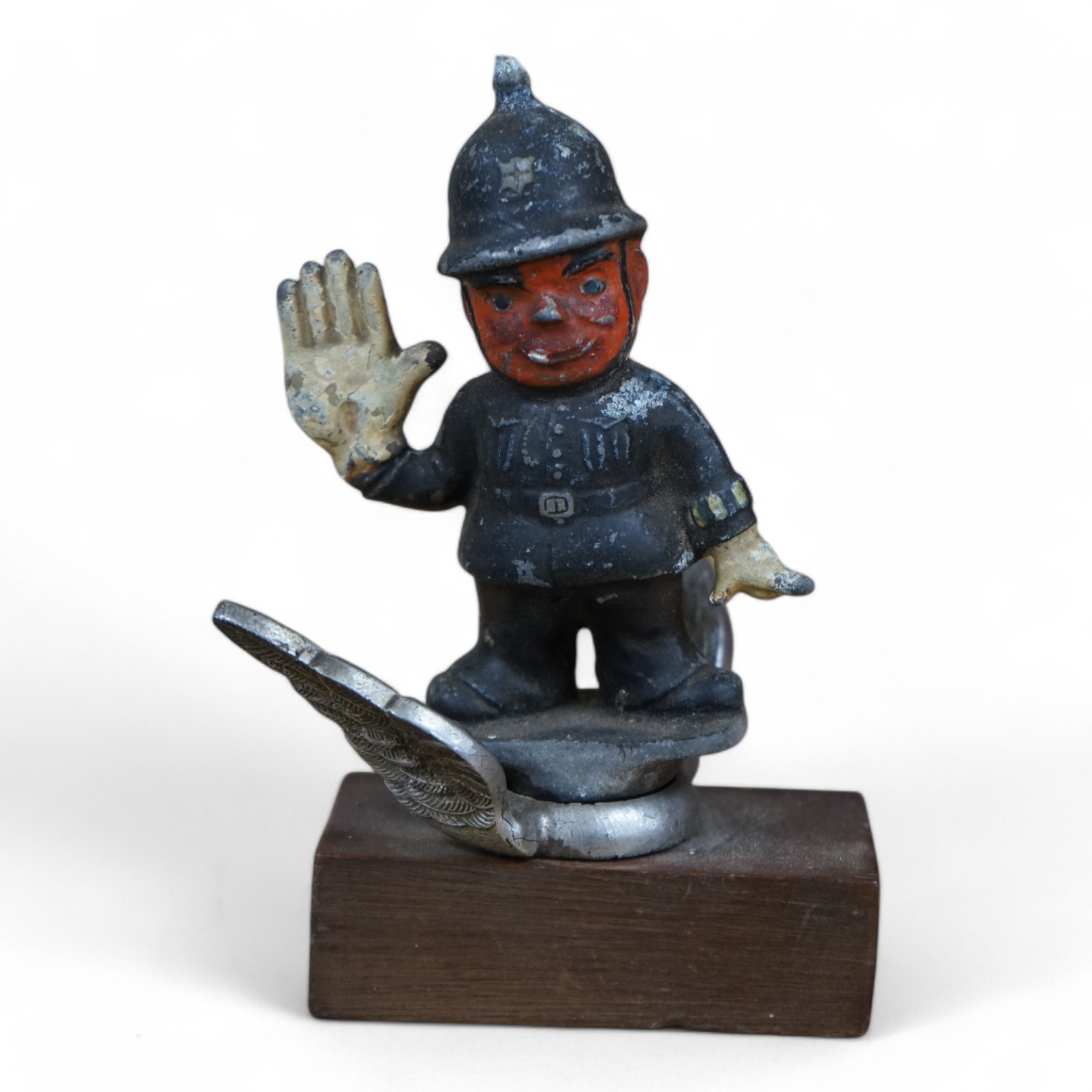 A cold painted cast aluminium car mascot in the form of a caricature of a policeman on associated chromed wings, mounted on a wooden block, policeman 11cm high. Condition - poor to fair.